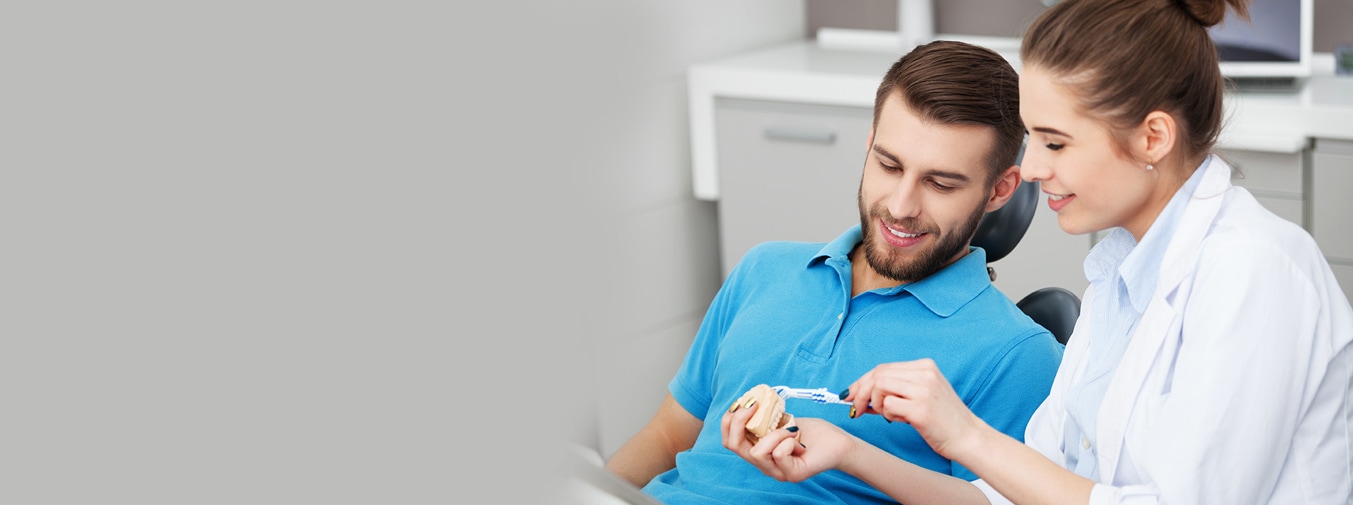 photo of patient watching the dental hygienist as she holds a mouth model and brush, the Advanced care plan icon (white square that has the Colgate logo inside a red rectangle) placed bottom right